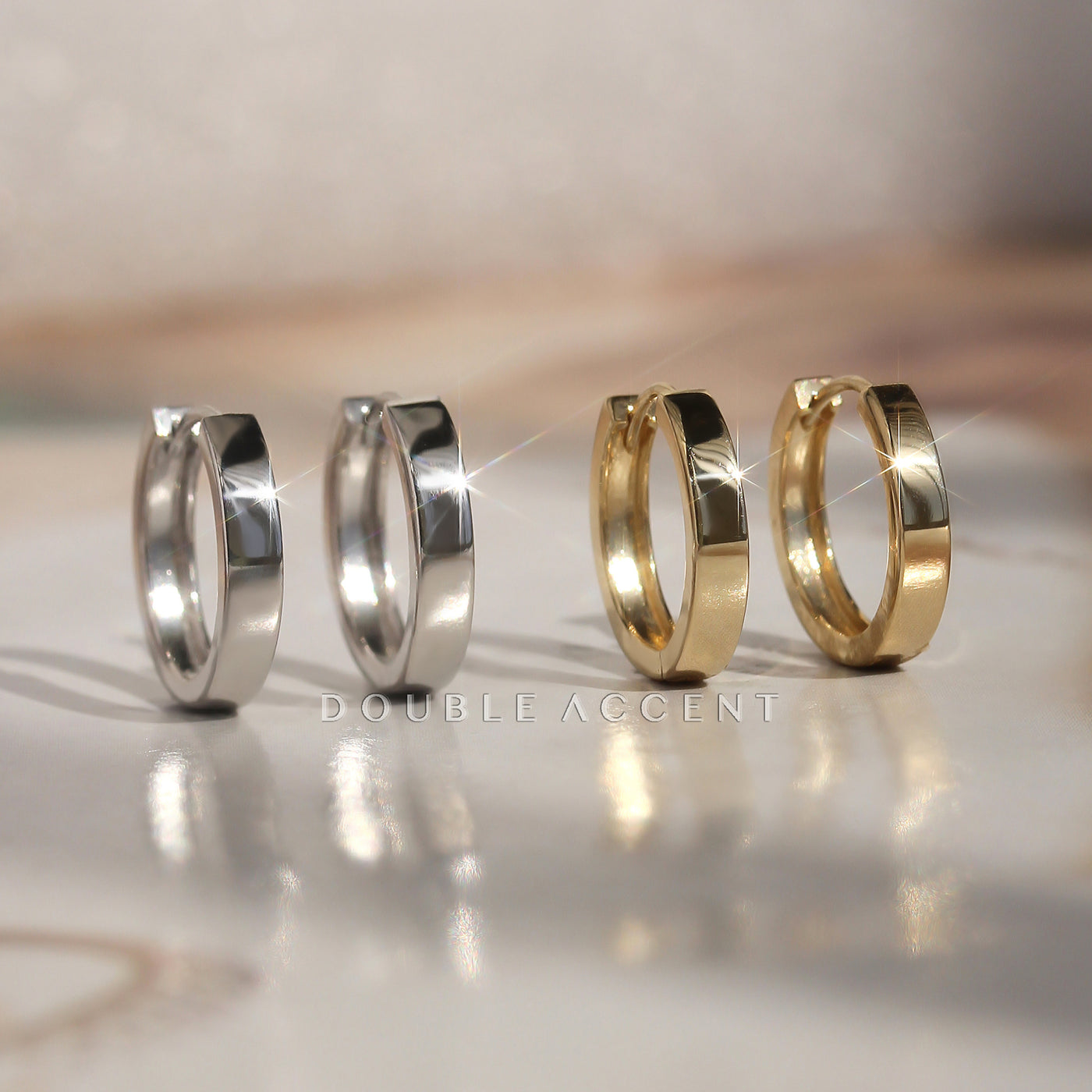 Two pairs of hoop earrings, one silver and one gold, on a reflective surface with 'Double Accent' branding.